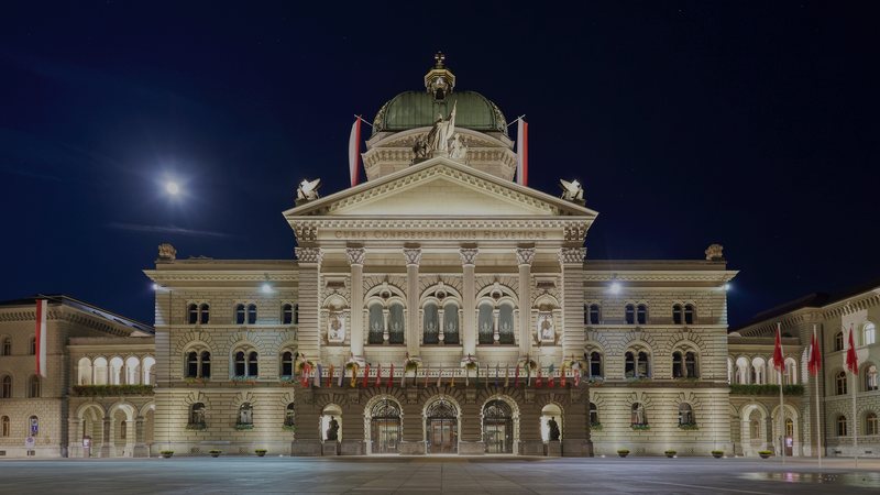 Foto: beleuchtetes Bundeshaus in Bern bei Nacht, mit Schweizer Fahnen am Bundesplatz
